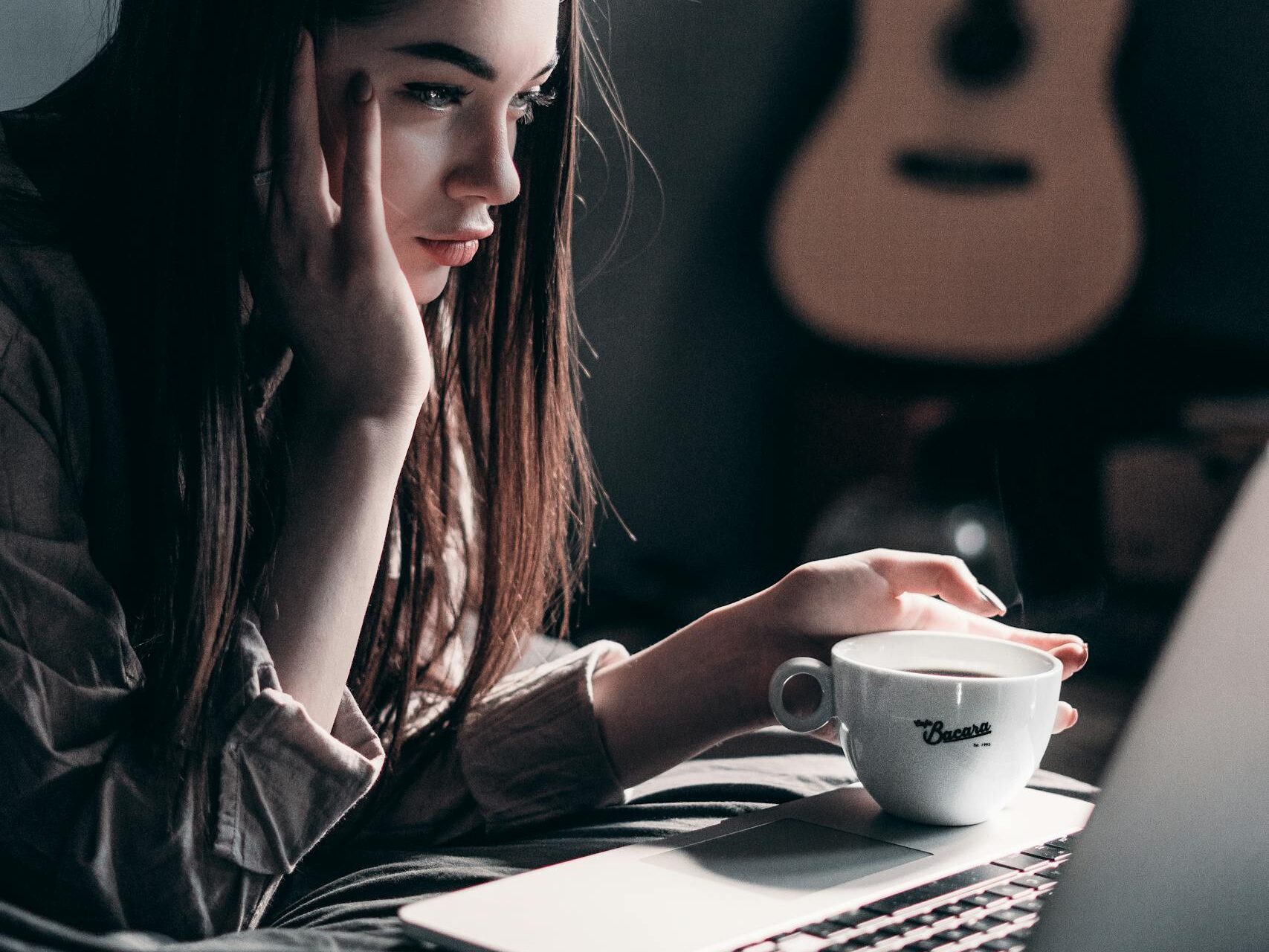 Woman lying on bed, focused on laptop with a coffee cup in hand, embracing remote work lifestyle.